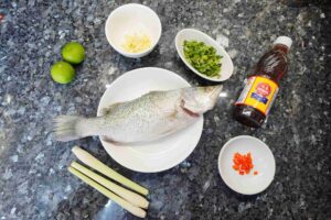 Thai-style steam fish recipe - all ingredients are prepared, fish in the middle, fresh lemongrass, lime, garlic, chopped coriander, fish sauce and chili are laying around the fish.